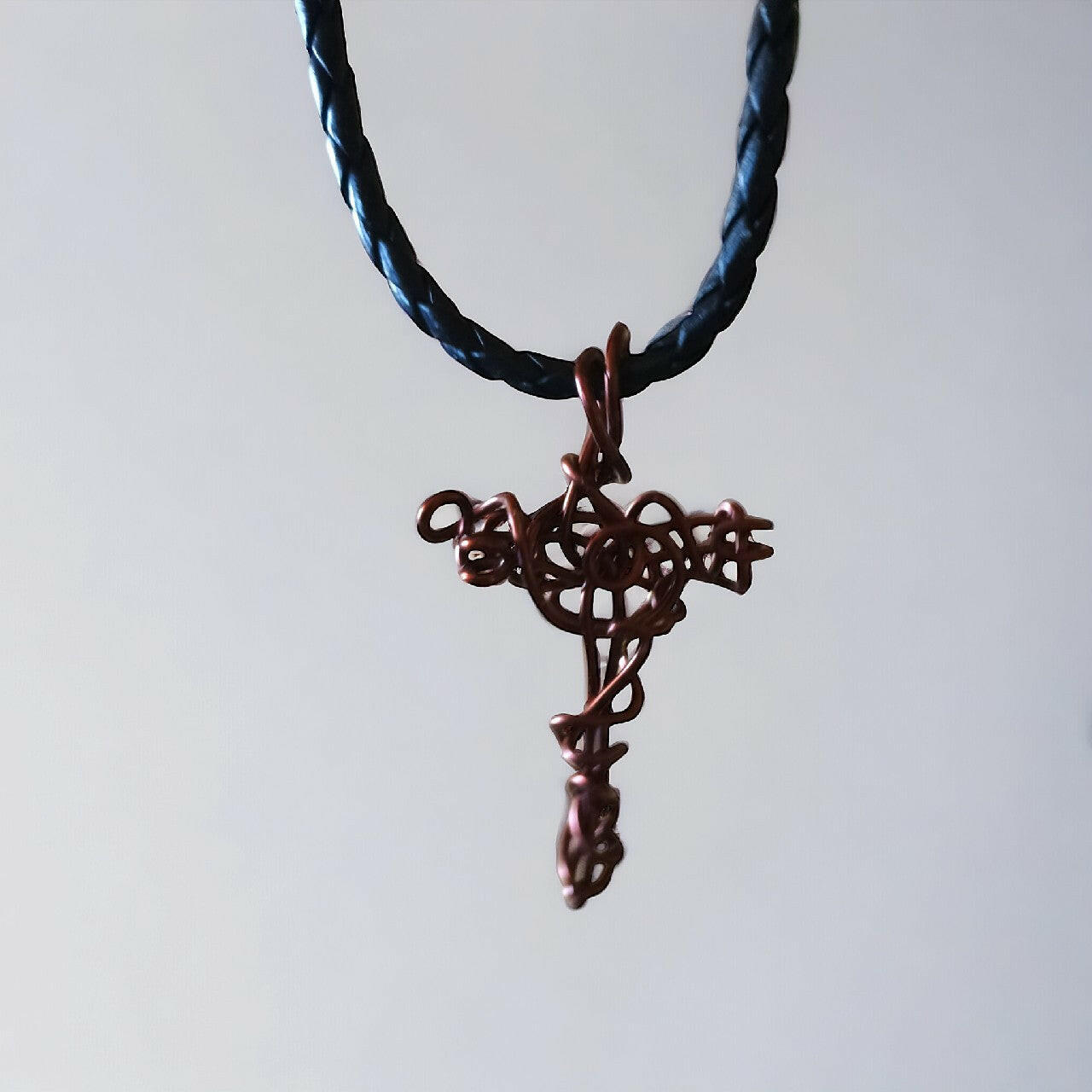 Ręcznie formowany wisiorek w kształcie krzyża z czystej miedzi, z ozdobnymi detalami wykonanymi techniką wire wrapping. Zawieszony na trwałym, plecionym rzemieniu w kolorze czarnym z mocnym zapięciem