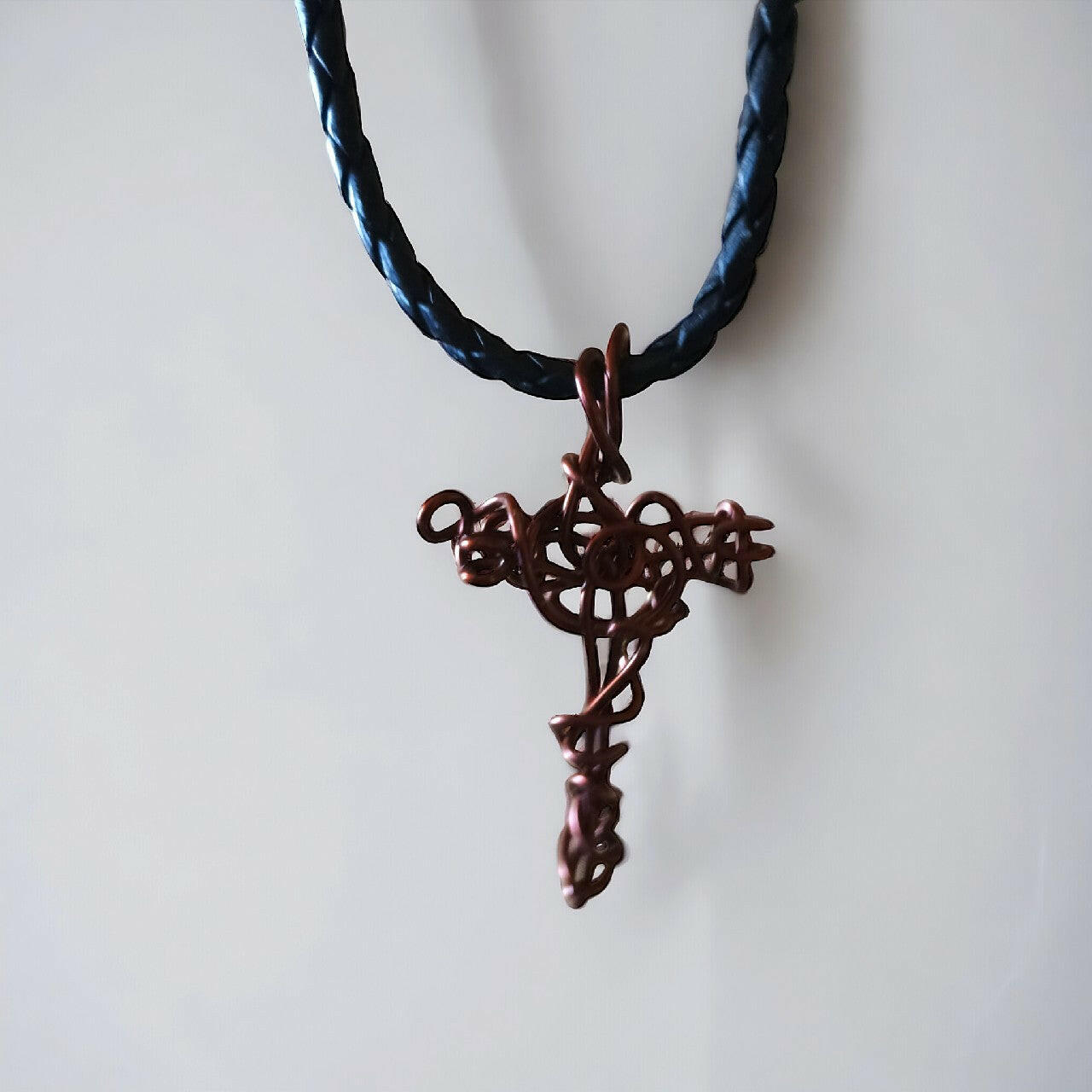 Artystyczny wisiorek przedstawiający krzyżyk z czystej miedzi, z unikalną strukturą uzyskaną dzięki technice wire wrapping. Zawieszony na eleganckim, plecionym rzemieniu w kolorze czarnym z solidnym zapięciem.