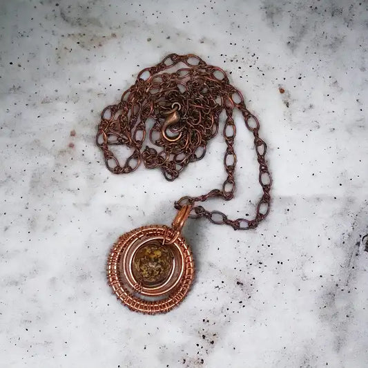 Rustykalna elegancja z akcentem natury: Okrągły wisiorek, w którym surowy, lecz fascynujący kamień jaspisu jest centralnym punktem, otoczonym misterną, miedzianą konstrukcją. Miedziany łańcuszek idealnie dopełnia ten inspirowany naturą dodatek, nadając mu charakterystyczny, rustykalny styl.