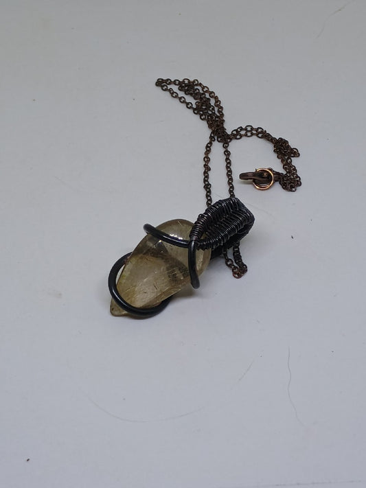 Zbliżenie unikalnego wisiorka z igiełkami rutylu zatopionymi w kwarcu, oprawionego w oksydowaną miedź. Misterna praca wire wrapping podkreśla naturalne piękno kamienia